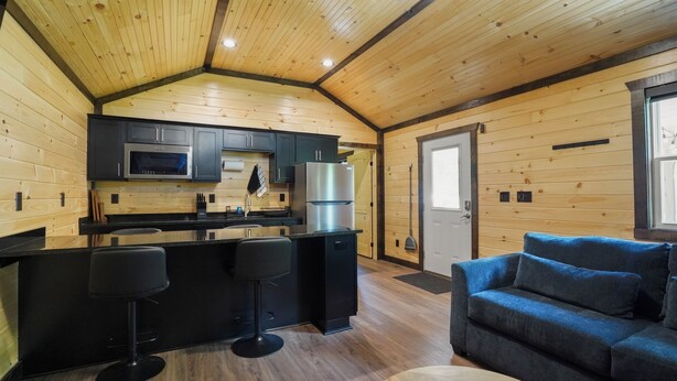 A wooden cabin interior with a kitchen, bar stools, a sofa, and a remote control on a table. - Route 66 Retreat (Somerset)