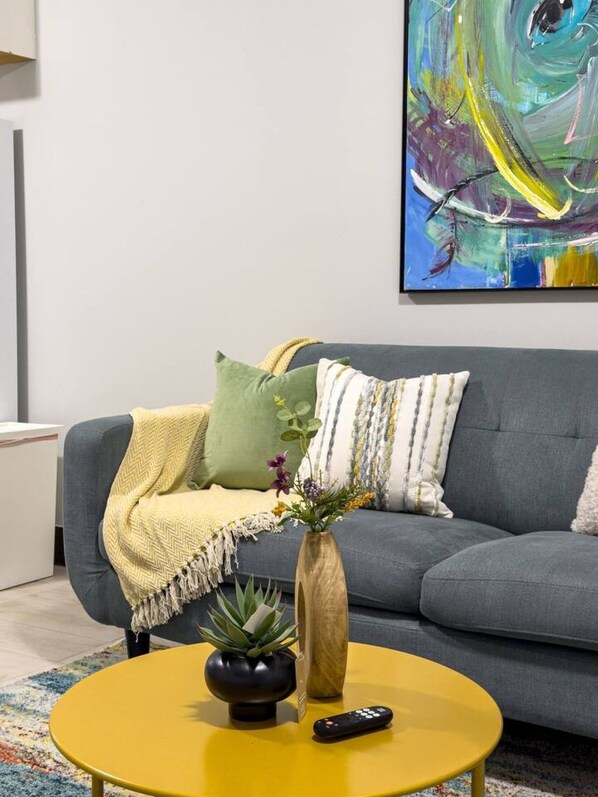 Un salon avec un canapé gris, une table basse jaune, un vase avec des fleurs et une t élécommande.