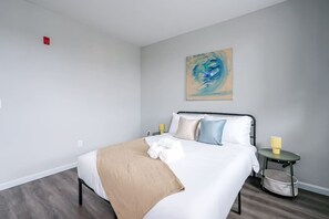 Une chambre d’hôtel avec un lit, un tableau au mur, une table de chevet avec une lampe et une petite poubelle.
