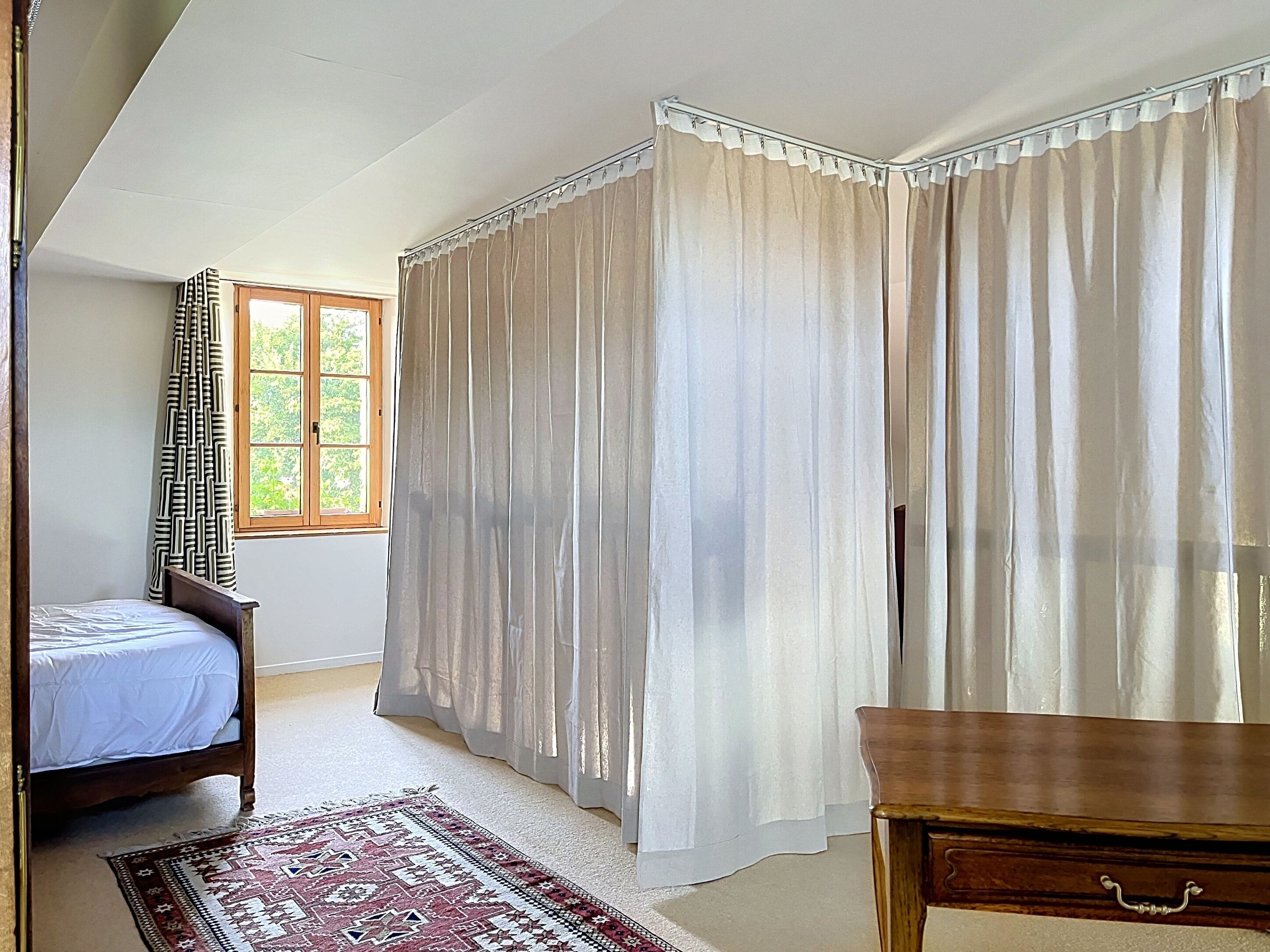 A bedroom with a bed, a window, curtains, a wooden nightstand, and a patterned rug.