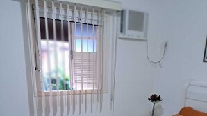 Ein Schlafzimmer mit einem Bett, einem Fenster mit Jalousien, einem Nachttisch mit einer Vase und einer Wand-Klimaanlage.