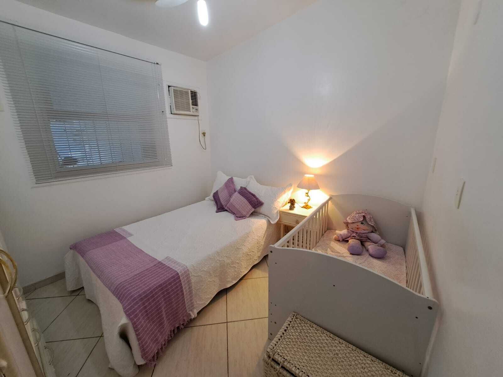 A small, clean room with a bed, a nightstand, a crib, a window with blinds, and an air conditioning unit.