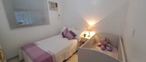 A small, clean room with a bed, a nightstand, a crib, a window with blinds, and an air conditioning unit.