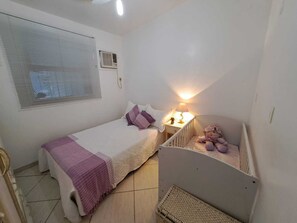 A small, clean room with a bed, a nightstand, a crib, a window with blinds, and an air conditioning unit. - Family home in a gated community. FAMILY ENVIRONMENT. (Angra dos Reis)