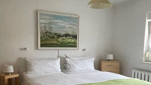 Ett sovrum med en säng, ett sängbord, en liten avlastningsyta, en lampa, ett element och en tavla på väggen.