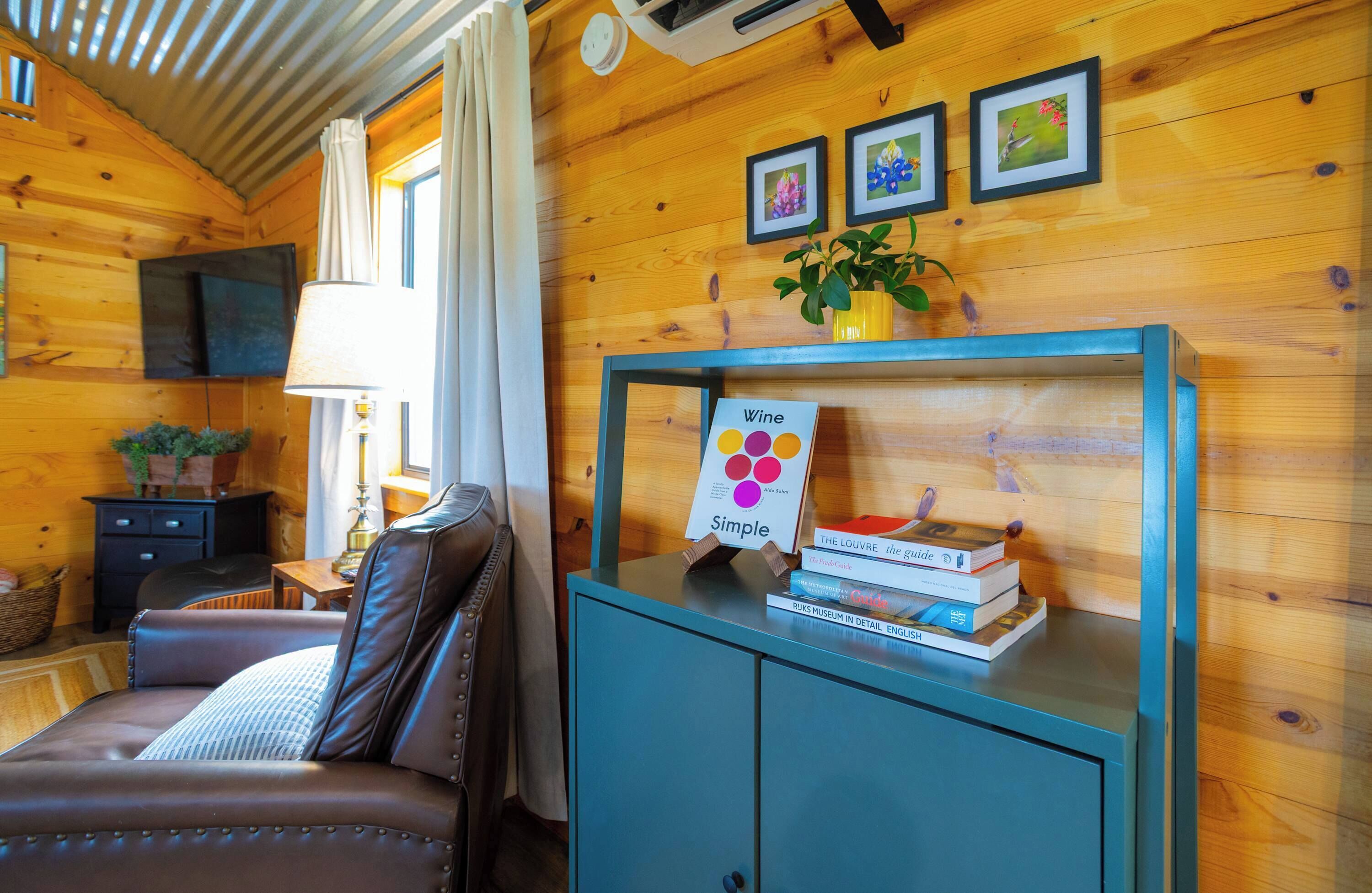 A wooden cabin interior with a sofa, a table with books, a wall-mounted TV, and framed pictures.