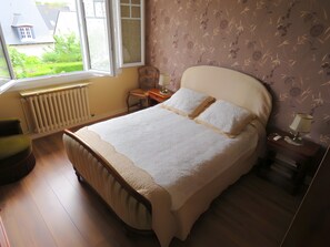Een slaapkamer met een bed, een stoel, een nachtkastje, een lamp en een raam met uitzicht op een huis en groen.