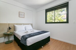 Un dormitorio con cama, mesita de noche con lámpara, ventana con vista y un cuadro en la pared.