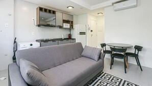 Un espace de vie moderne comprenant un canapé gris, une table à manger avec des chaises, un coin cuisine avec un four à micro-ondes et un réfrigérateur, ainsi qu’un appareil de climatisation fixé au plafond.
