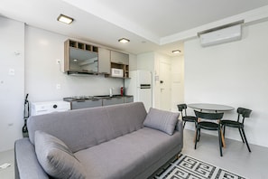 Ein stilvolles Wohnzimmer mit grauem Sofa, einem Esstisch mit Stühlen, einer Küchenzeile mit Mikrowelle und Kühlschrank sowie einer Decken-Klimaanlage.