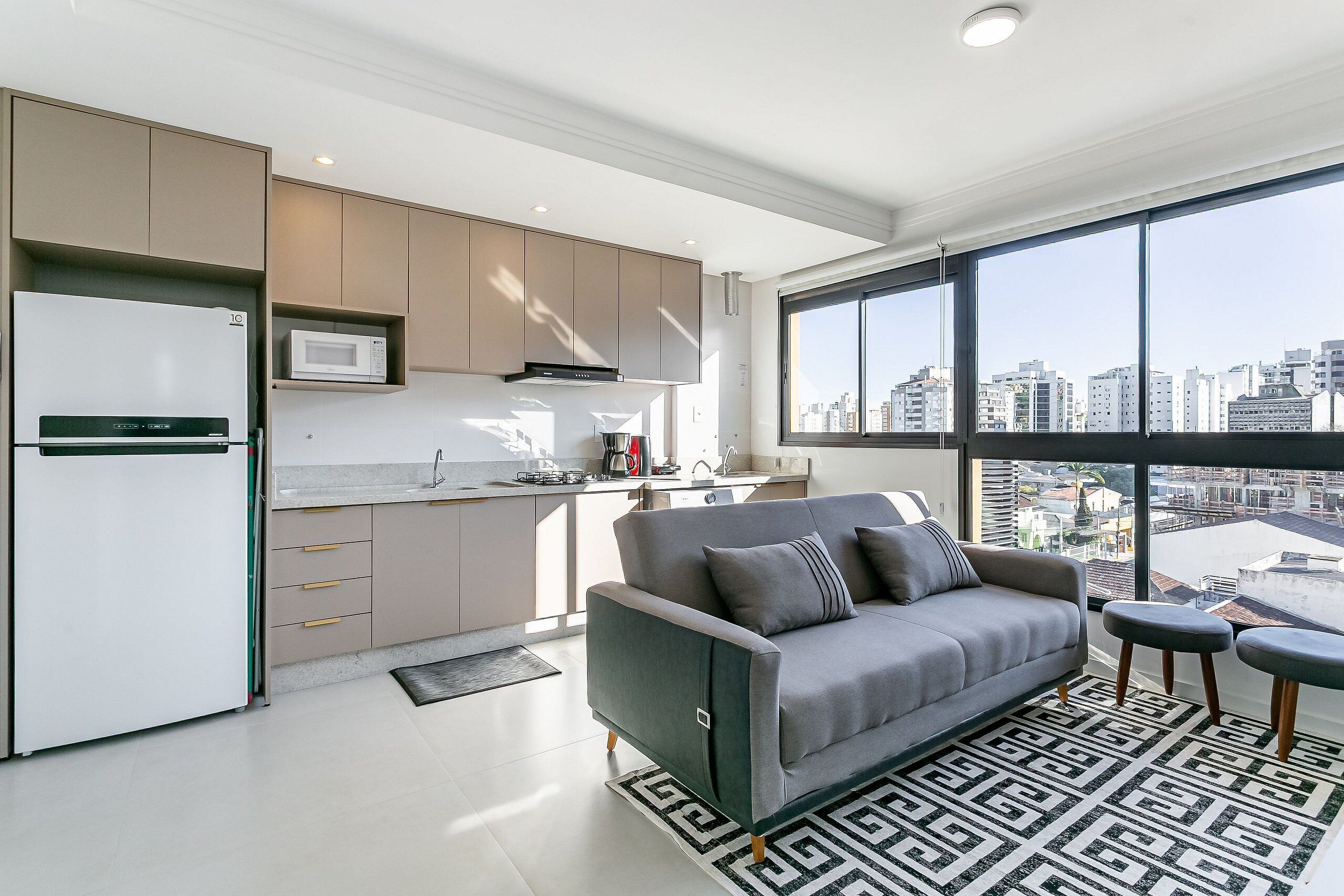 Eine moderne Küche mit integriertem Mikrowellenherd, einem Kühlschrank und einer Kaffeemaschine. Ein graues Sofa mit Kissen und ein gemustertes Teppich sorgen für Gemütlichkeit. Ein großes Fenster bietet Blick auf die Stadtlandschaft.