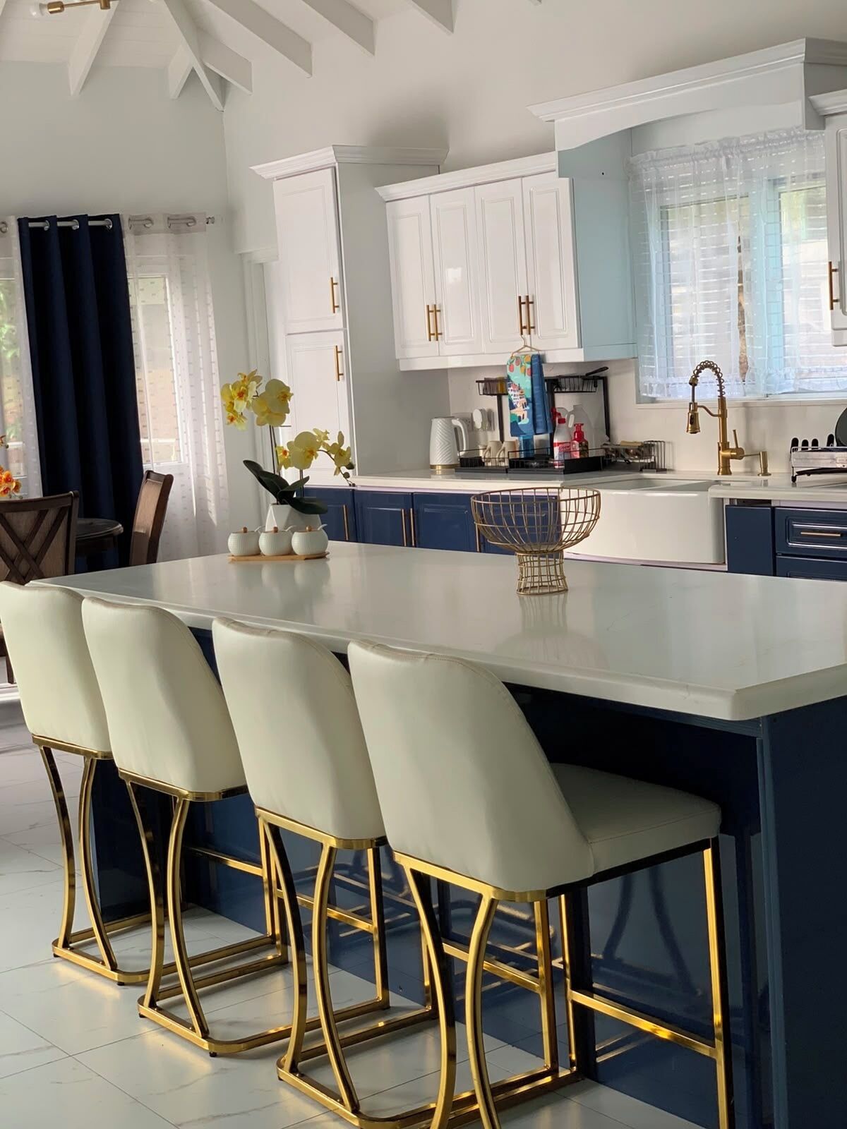 A modern kitchen with white cabinets, a central island with bar stools, and a window with sheer curtains.