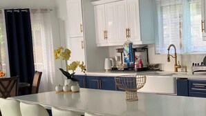 Une cuisine moderne avec des meubles blancs, un îlot central doté de tabourets de bar et une fenêtre ornée de rideaux légers.