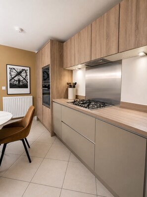 A modern kitchen with wooden cabinets, a gas stove, and a dining area with a chair. - Delightful 3-bedroom house in charming County Durham perfect for families. (Chilton)