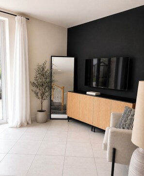 A modern living room with a large flat-screen TV mounted on a black wall, a wooden console table, a potted plant, and a sofa with striped cushions. - Delightful 3-bedroom house in charming County Durham perfect for families. (Chilton)