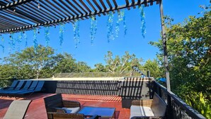 A covered outdoor seating area with lounge chairs and a table, surrounded by trees and a clear blue sky. - Cuatro Elementos By Gestores Studio (Tulum)