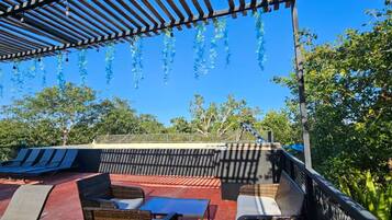 Un espace extérieur aménagé, abrité et doté de chaises longues et d’une table, entouré d’arbres et surplombé d’un ciel bleu dégagé.