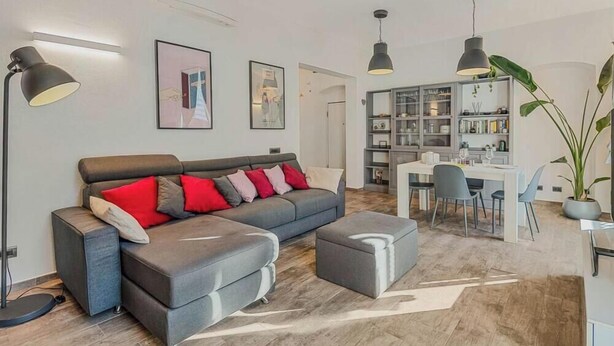 A modern living room with a grey sectional sofa, a dining area with a white table and chairs, and a bookshelf filled with books and decorative items. - Graffiti House Luxury Apartment (La Spezia)