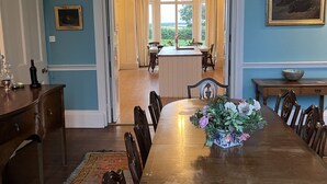 Een eetkamer met een grote houten tafel, stoelen, een bloevas en uitzicht door een deuropening naar een keuken.