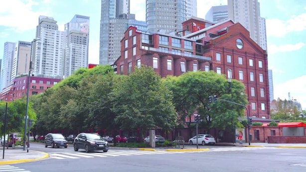 A cityscape with tall buildings, a red brick building with a circular window, and a crosswalk. - Hestia en Arenales 2900 (Buenos Aires)