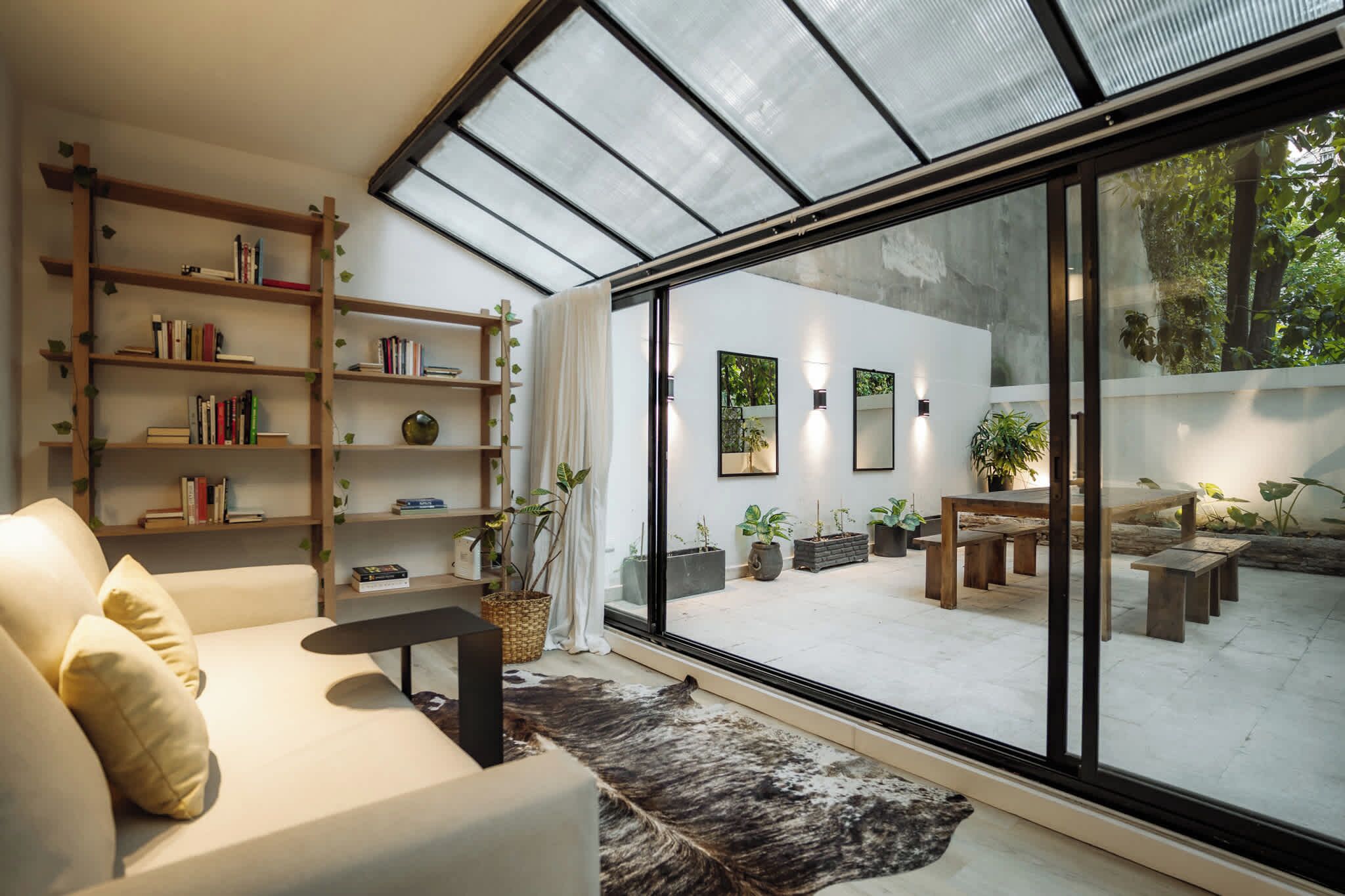 A modern living room with a sofa, a bookshelf, and a large glass door leading to an outdoor patio with plants and seating.
