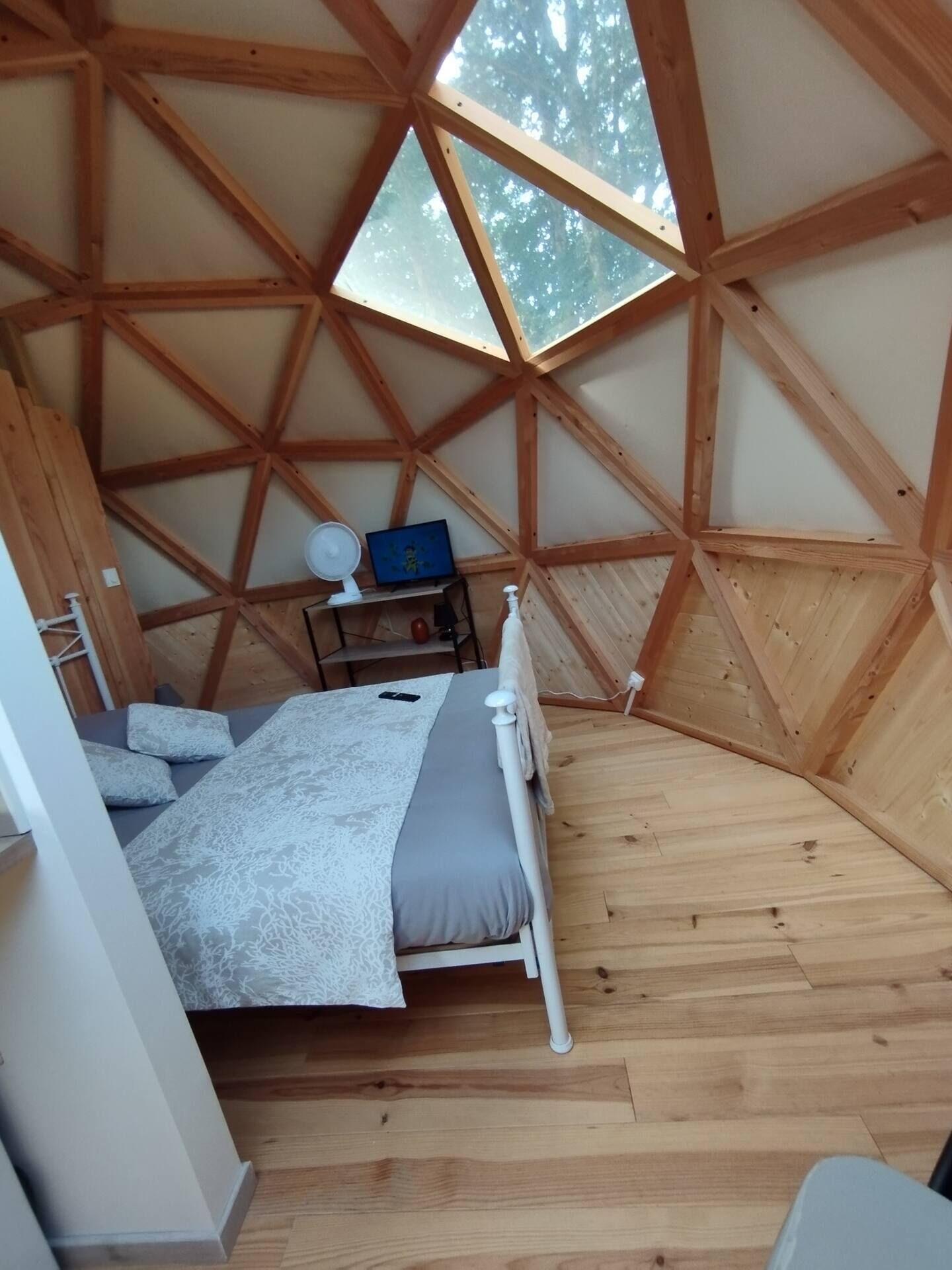 A room with a sloped ceiling, wooden beams, a bed with a white headboard, a bedside table with a lamp, and a window with a view of trees.