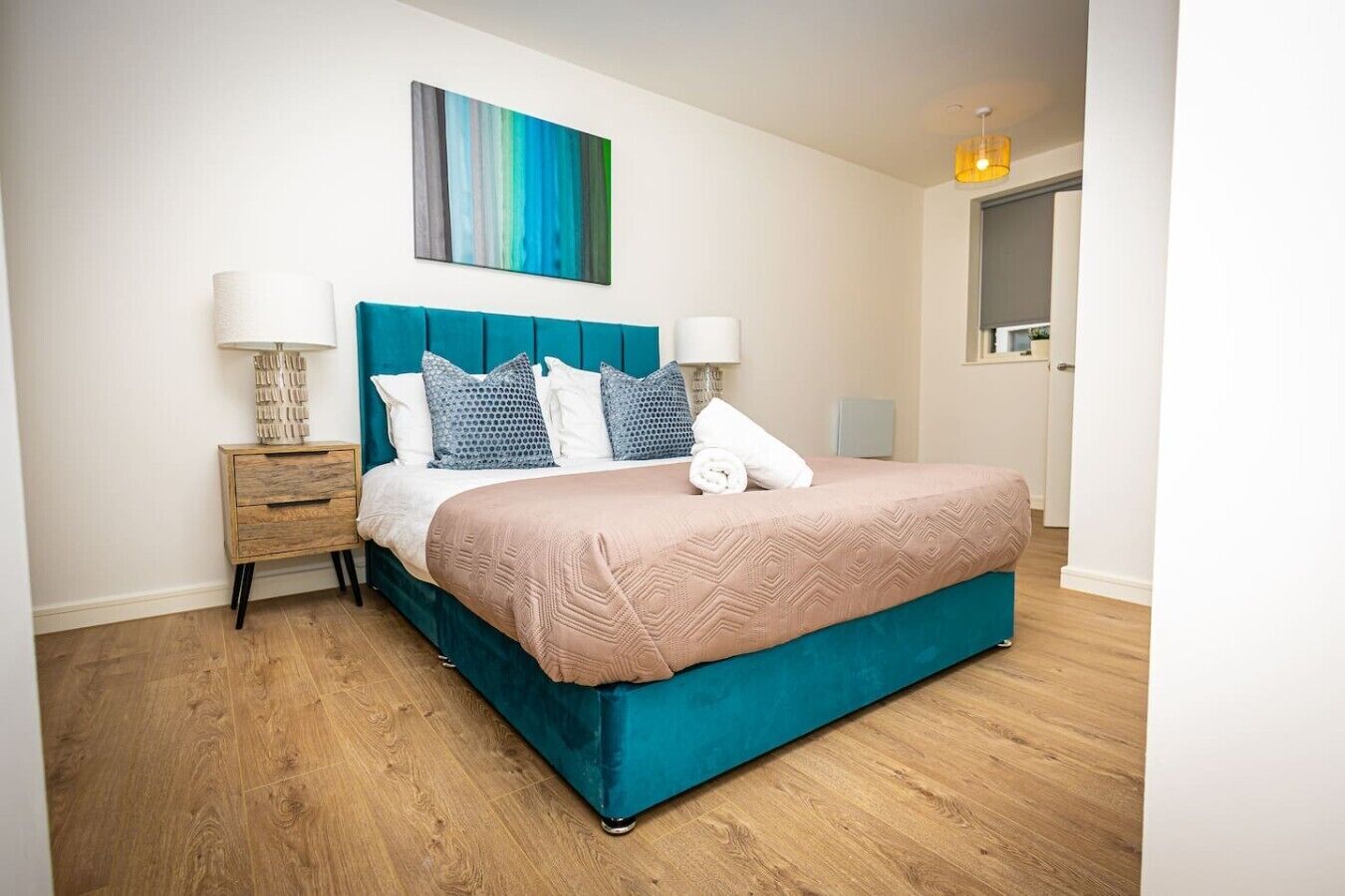 A modern bedroom with a teal upholstered bed, a wooden nightstand, and a painting on the wall.