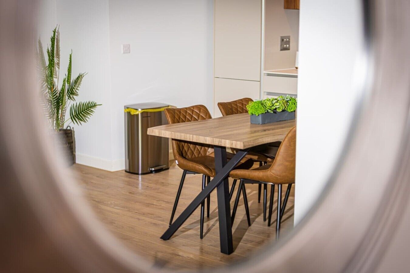 A modern dining area with a wooden table, leather chairs, a potted plant, and a trash bin.