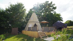 Une structure triangulaire en forme de tente, avec un escalier en bois menant à son sommet, entourée de verdure et d’un petit jardin.