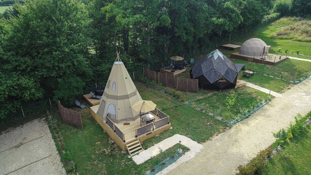 Un terrain de camping comprenant une tente conique, une structure en forme de dôme et une terrasse en bois avec des meubles d’extérieur.