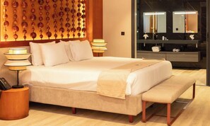 Quarto de hotel com uma cama grande, luminárias de cabeceira, um banco, um espelho fixado na parede e uma cômoda com espelho.