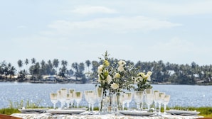 A table set with glassware and plates, a floral centerpiece, and a scenic lake view. - Escapade Hotel (Assinie)