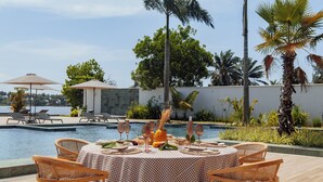 A poolside dining area with a checkered tablecloth, wicker chairs, and a view of the pool and palm trees. - Escapade Hotel (Assinie)