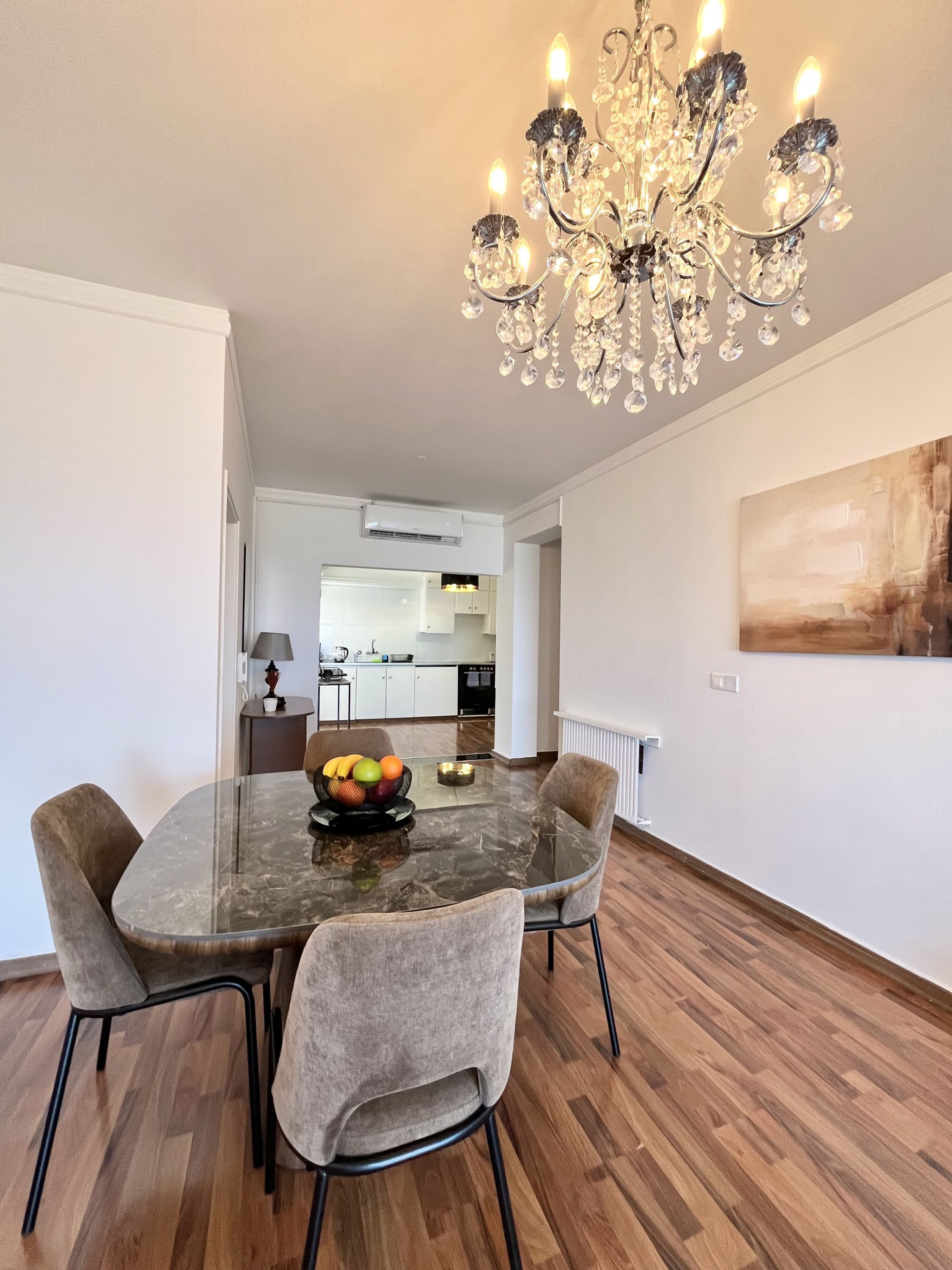 Een eethoek met een glazen tafel, vier stoelen, een kroonluchter, een fruitschaal en een keuken op de achtergrond.