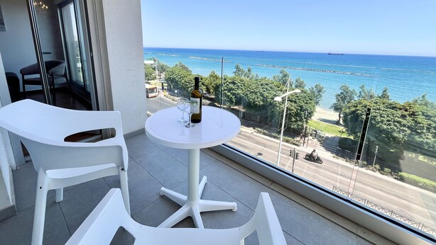 A balcony with a round table, two white chairs, a bottle of wine, and a view of the sea and greenery. - 159 LIM - Stay Sea Pearl Apartment (Limassol)