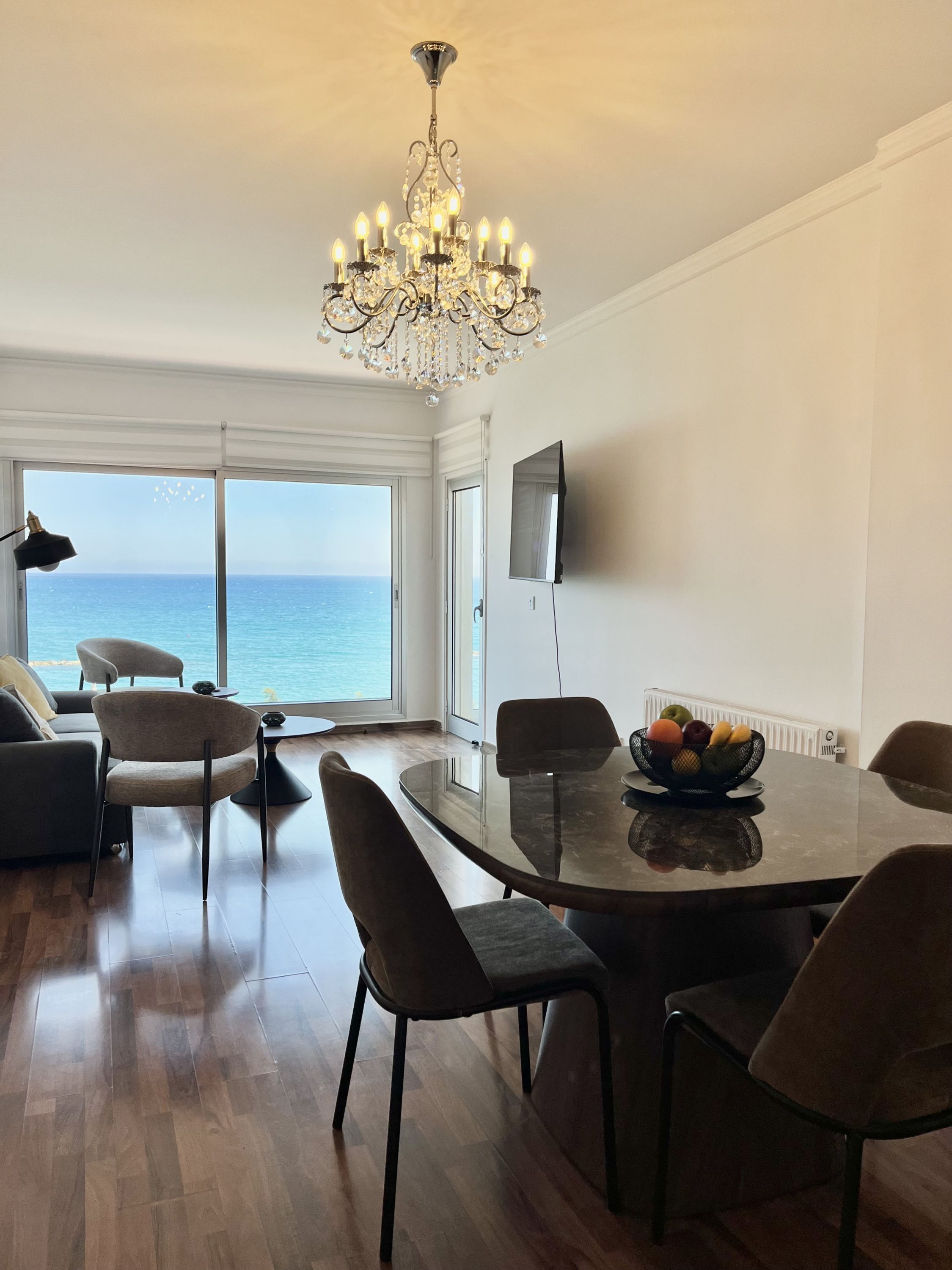A modern dining area with a chandelier, a dining table with chairs, a fruit bowl, and a view of the sea through large windows.