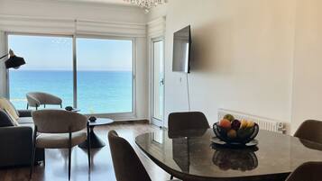 A modern dining area with a chandelier, a dining table with chairs, a fruit bowl, and a view of the sea through large windows.