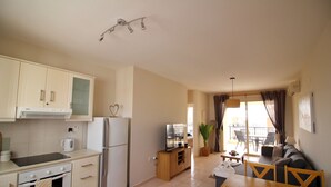 Et kompakt køkken med moderne hårde hvidevarer, et spiseområde med et træbord og stole, samt en stue med en sofa og et stort vindue.