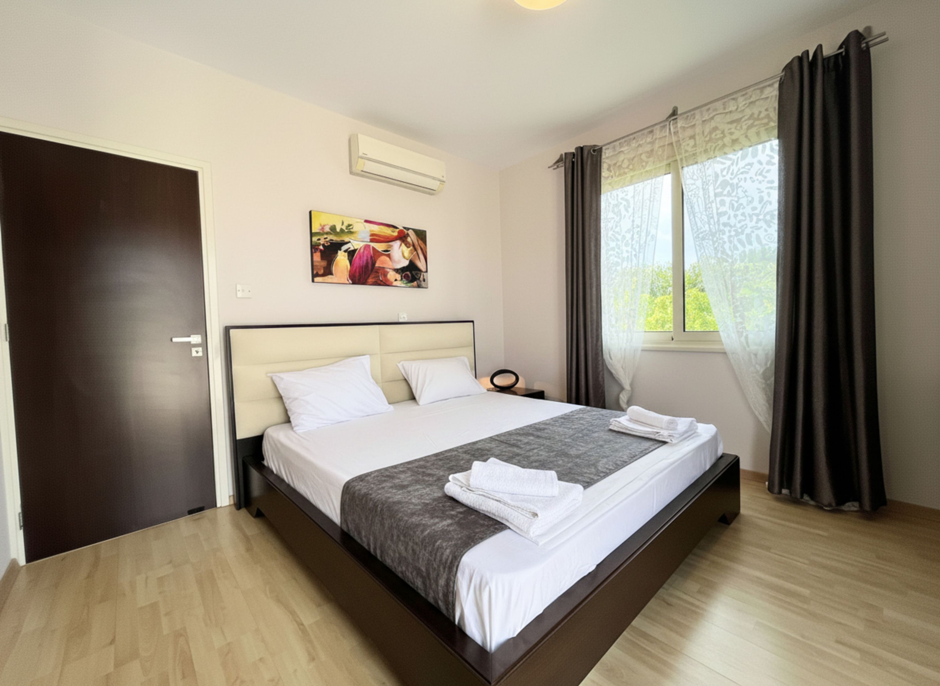 Habitación de hotel con una cama, un cuadro en la pared, aire acondicionado, una ventana con cortinas y una puerta.