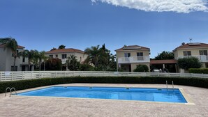 Una piscina con un borde de baldosas, rodeada por una valla blanca, y casas con balcones al fondo.