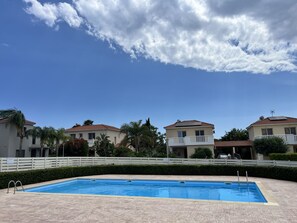 Una piscina con un borde de baldosas, rodeada por una valla blanca, y casas con balcones al fondo.