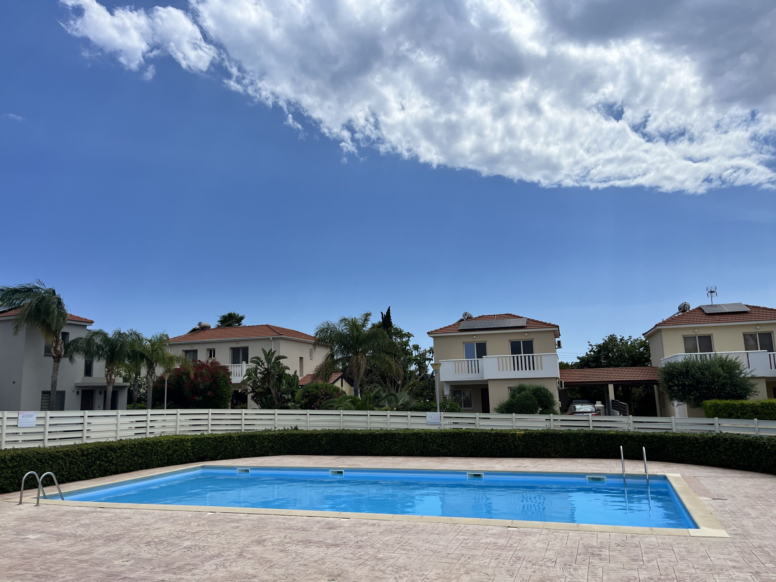 Una piscina con un borde de baldosas, rodeada por una valla blanca, y casas con balcones al fondo.