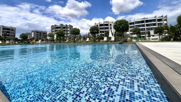 Una piscina con piso de baldosas, rodeada por una terraza de madera y sillas de descanso. Al fondo, edificios de varios pisos con balcones.