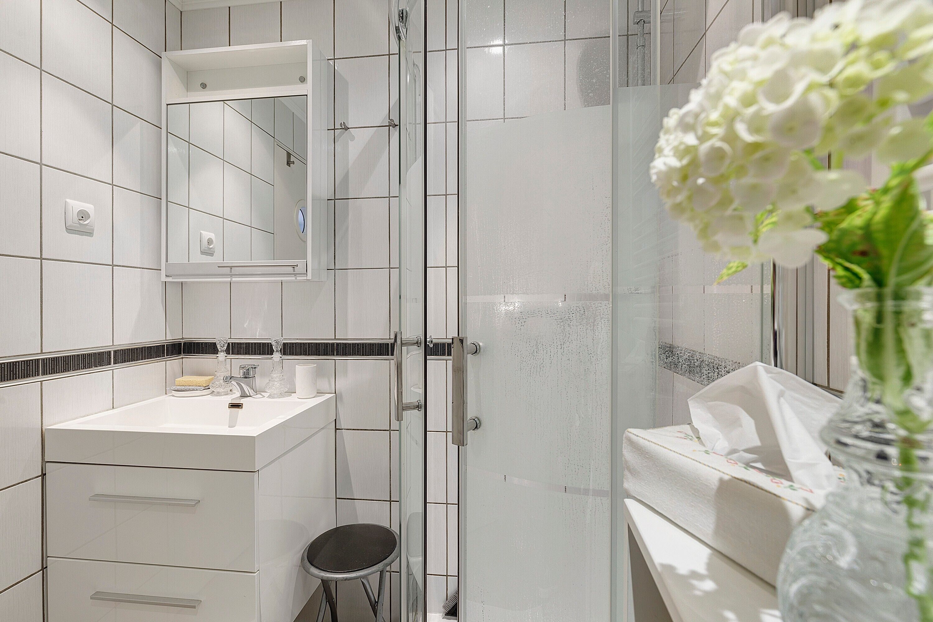 Une salle de bain avec une douche à parois de verre, un lavabo blanc avec un miroir et un vase contenant des fleurs blanches.