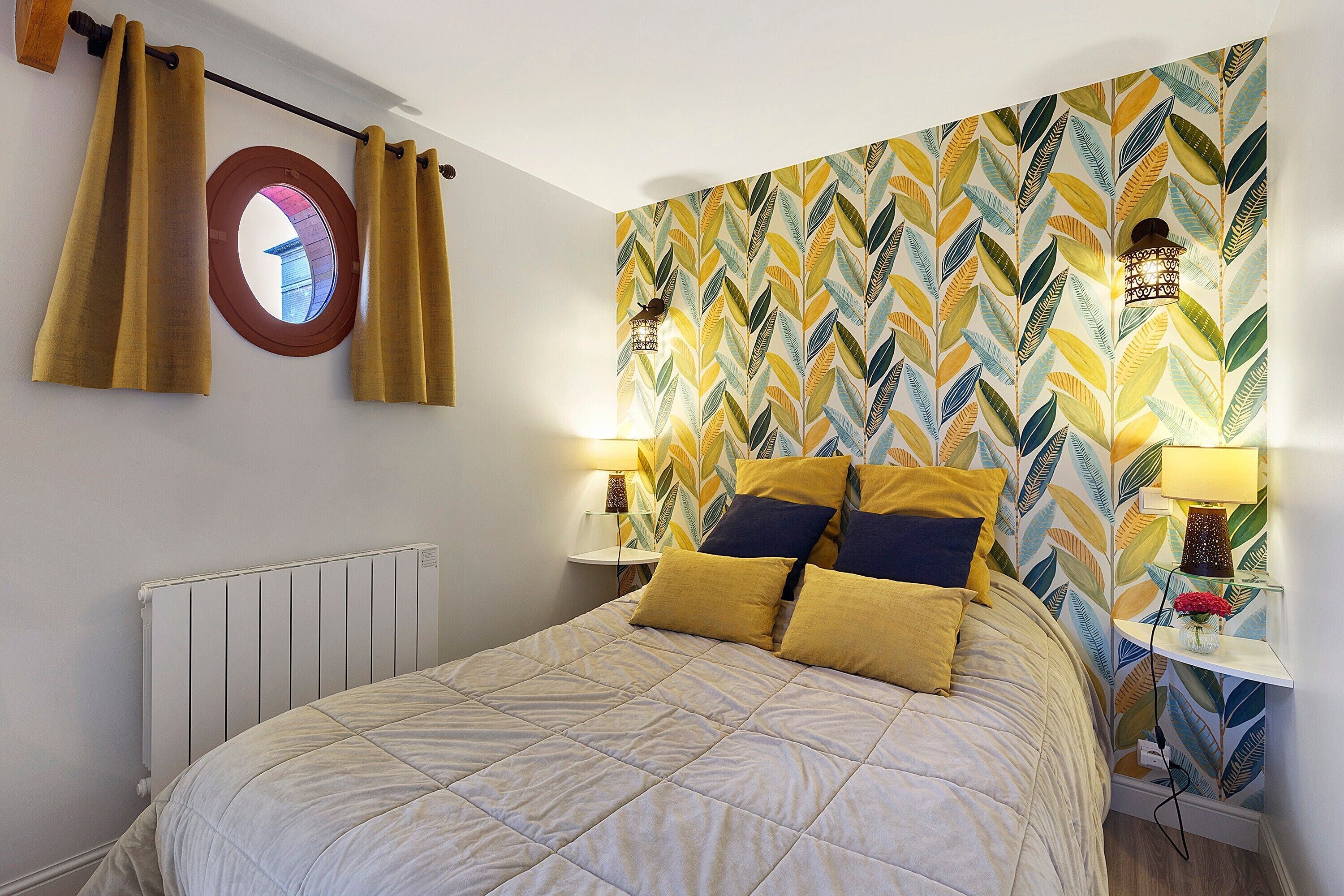 Quarto com cama, duas luminárias de cabeceira, radiador e janela com cortinas.