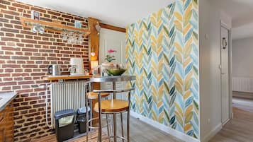 Une cuisine avec un mur en briques, une étagère en bois où sont rangés les ustensiles de cuisine, une table ronde avec des tabourets et un papier peint à motifs.