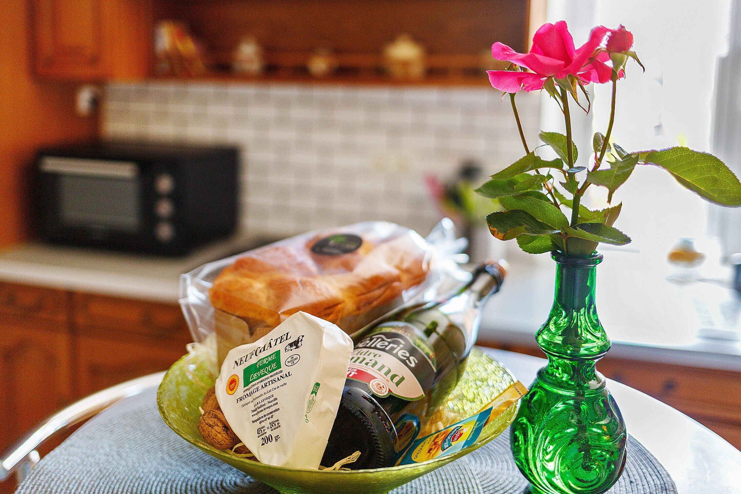A green glass bottle with a pink flower, a glass bowl with a baguette, and a bottle of wine.