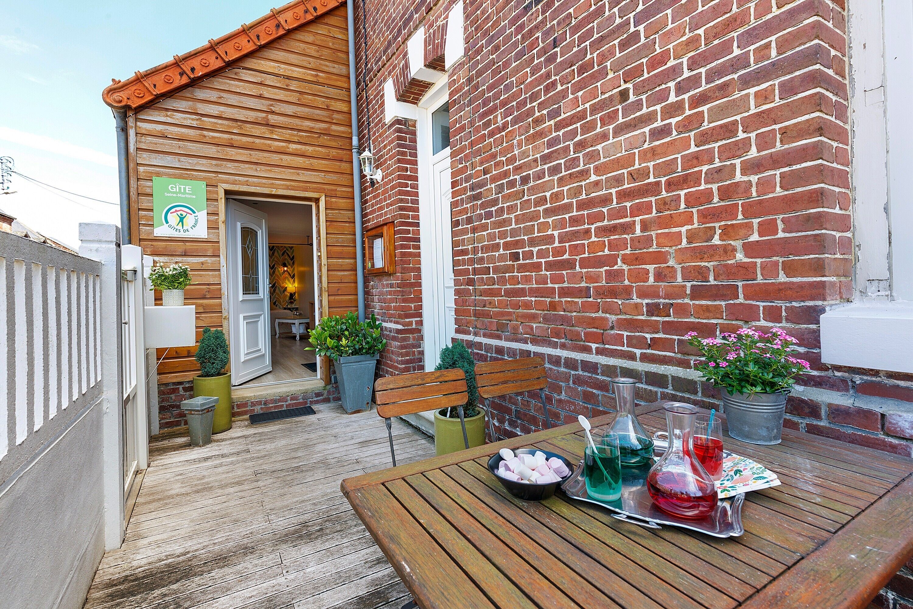 A wooden deck with a table set for dining, a brick wall, and a door leading to a room.