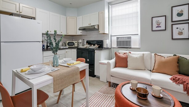 A compact kitchen with white cabinets, a dining table with chairs, a white sofa, and a window with blinds. - Lancaster Art Suite 4 - 2BR near Drexel & Penn (Philadelphia)