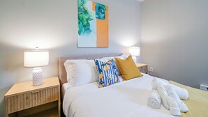 A neatly made bed with white linens, a blue and white striped pillow, and a mustard-colored pillow. There are two bedside lamps on wooden nightstands. A painting hangs above the bed. - Lancaster Art Suite 4 - 2BR near Drexel & Penn (Philadelphia)